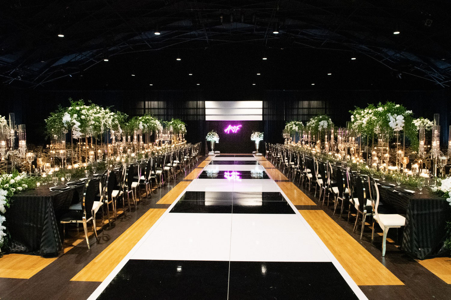 Elegant banquet setup with long tables adorned with greenery, candles, and a neon sign in a spacious venue.