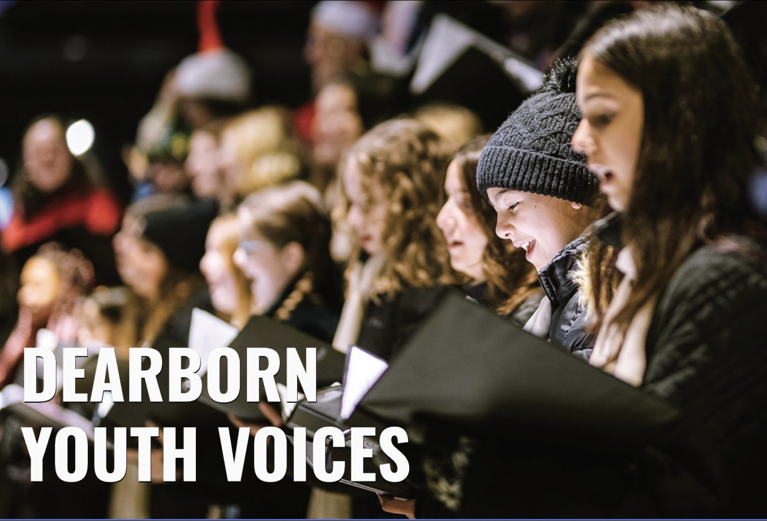 A group of young singers performing, with a focus on a smiling child in a black beanie, holding sheet music.